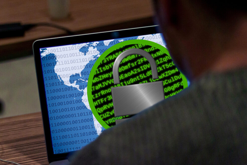Laptopbildschirm mit Weltkarte, Binärcode und großem Vorhängeschloss-Symbol vor grünem Codehintergrund, Person im Vordergrund.