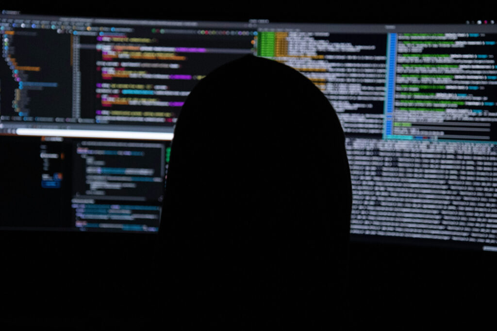Silhouette einer Person vor mehreren großflächigen Monitoren mit farbig hervorgehobener Programmier- und Konsolenausgabe.