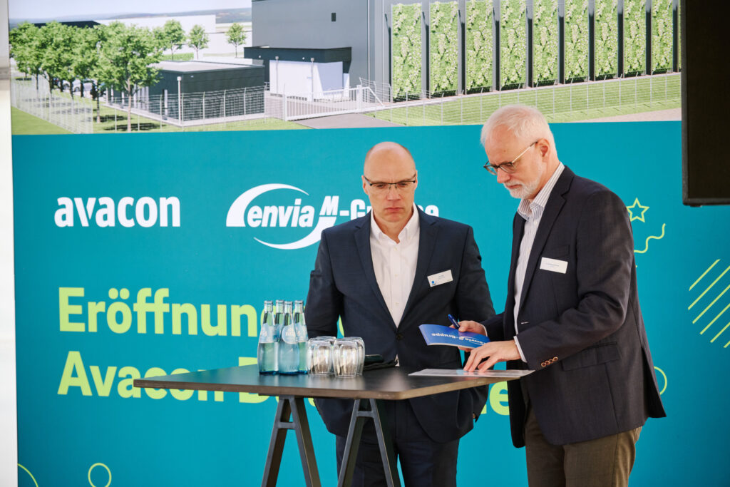 Zwei Männer in Anzügen prüfen Unterlagen an einem Stehtisch vor einer grünen Eventwand mit Logos und Wasserflaschen.