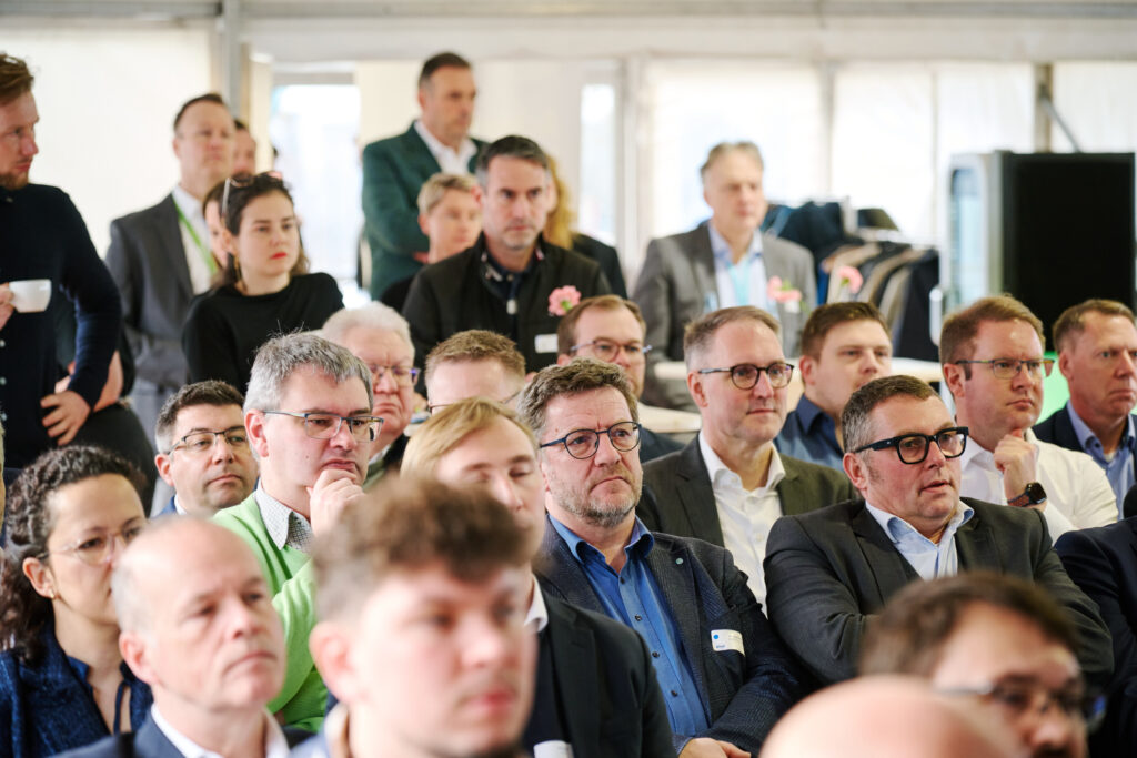Erwachsenes Publikum in Business‑Kleidung, aufmerksam sitzend bei einer Konferenzveranstaltung in einem Zeltraum.