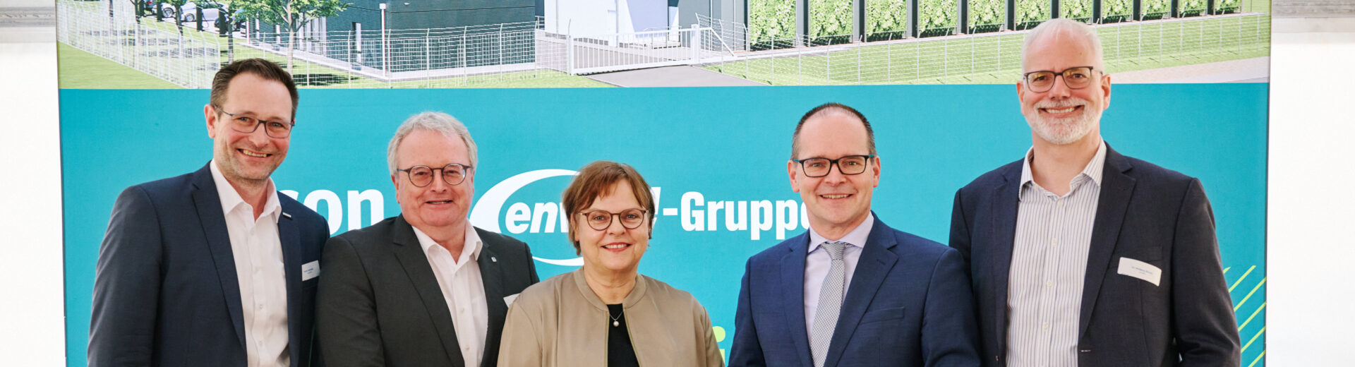 Fünf lächelnde Personen in Businesskleidung vor einem blau-grünen Messestand mit Gebäudezeichnung im Hintergrund.