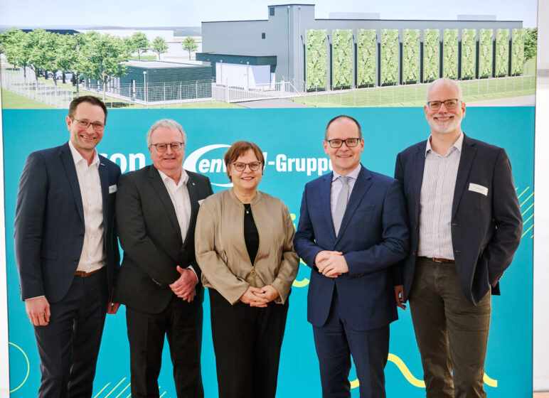 Fünf lächelnde Personen in Businesskleidung vor einem blau-grünen Messestand mit Gebäudezeichnung im Hintergrund.
