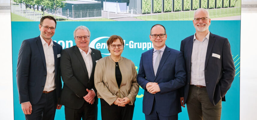 Fünf lächelnde Personen in Businesskleidung vor einem blau-grünen Messestand mit Gebäudezeichnung im Hintergrund.