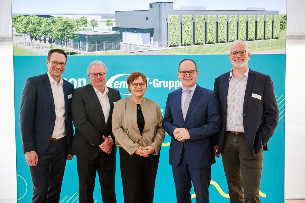 Fünf lächelnde Personen in Businesskleidung vor einem blau-grünen Messestand mit Gebäudezeichnung im Hintergrund.