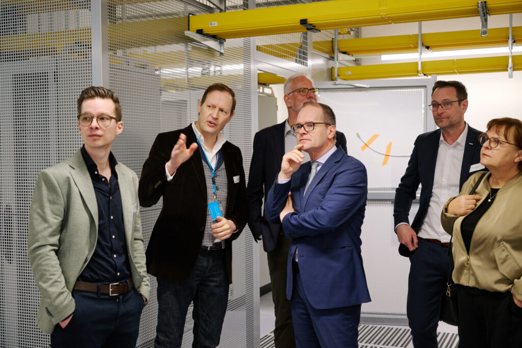 Gruppe von sechs Personen in Geschäftskleidung, eine erklärt etwas gestikulierend, andere hören aufmerksam in einer hellen Industrieumgebung.