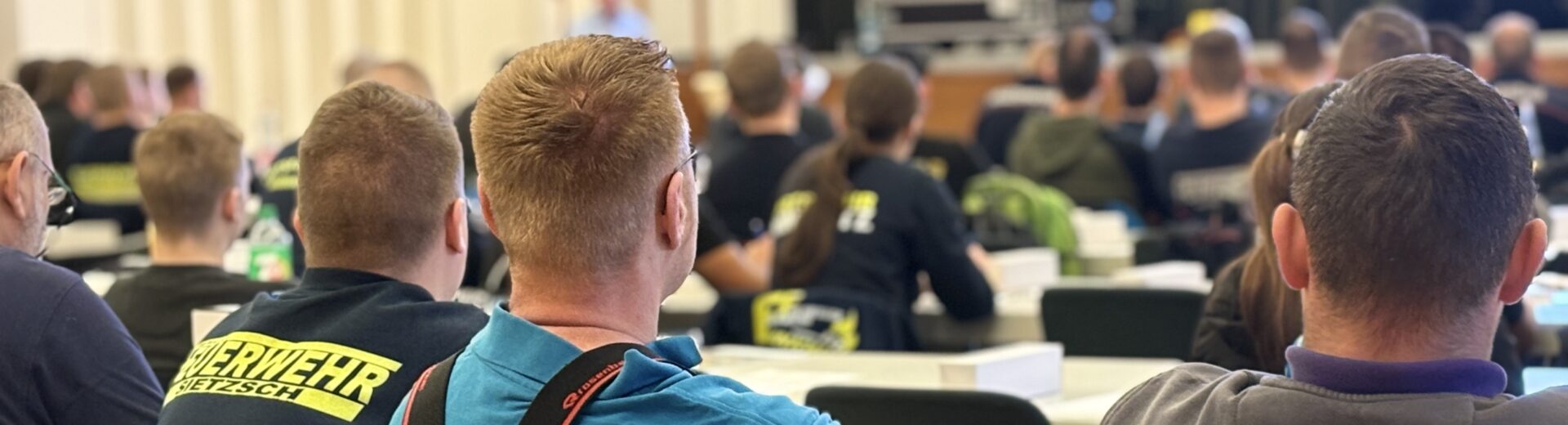 Mehrere Personen von hinten in einem Seminarraum, einige mit leuchtenden Feuerwehrjacken und Aufdrucken wie "Feuerwehr" und "GOLLMA".