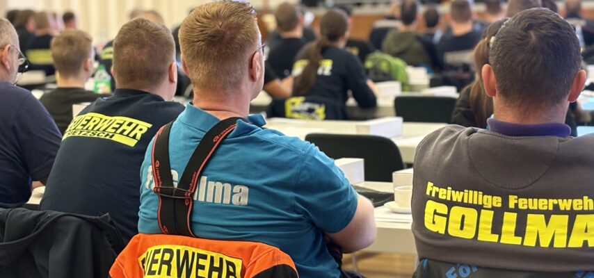 Mehrere Personen von hinten in einem Seminarraum, einige mit leuchtenden Feuerwehrjacken und Aufdrucken wie "Feuerwehr" und "GOLLMA".