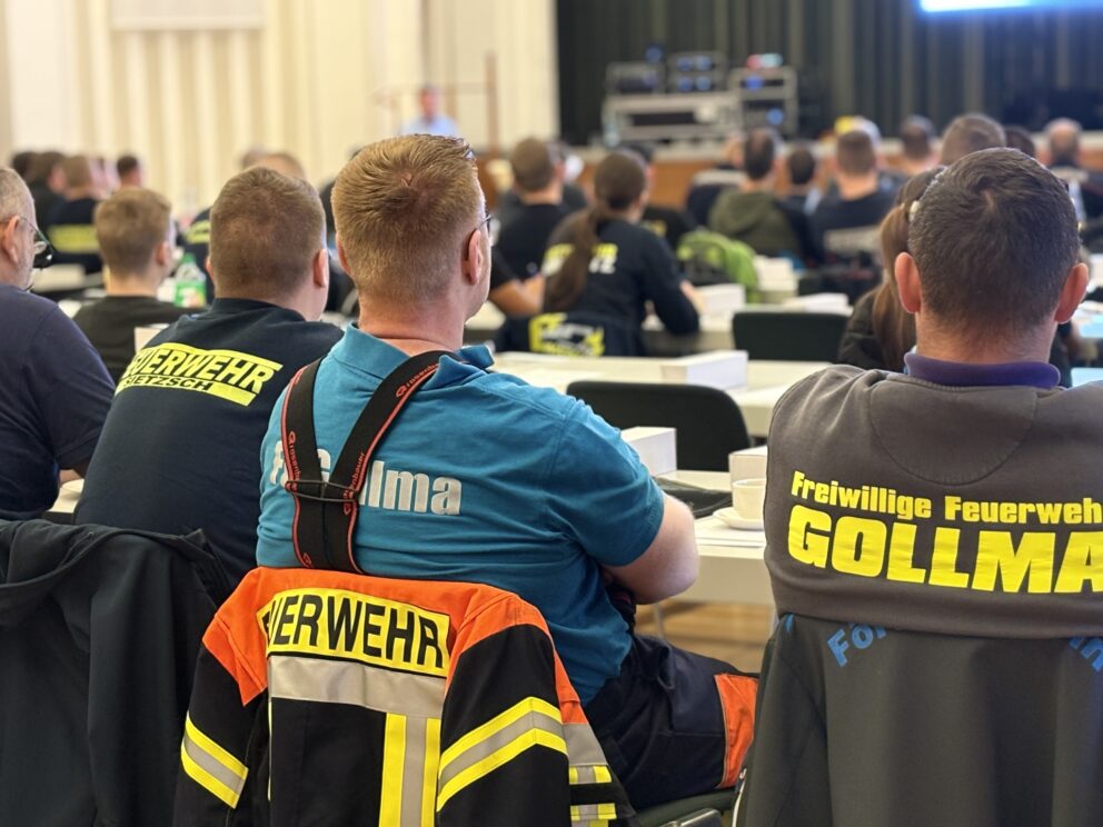 Mehrere Personen von hinten in einem Seminarraum, einige mit leuchtenden Feuerwehrjacken und Aufdrucken wie "Feuerwehr" und "GOLLMA".
