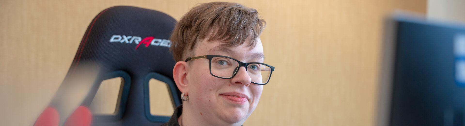 Person mit kurzen Haaren und Brille lächelt leicht vor einem Gaming-Stuhl, im Vordergrund unscharfer Monitor.