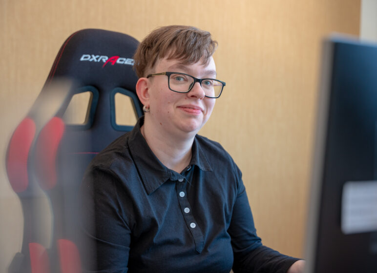Person mit kurzen Haaren und Brille lächelt leicht vor einem Gaming-Stuhl, im Vordergrund unscharfer Monitor.