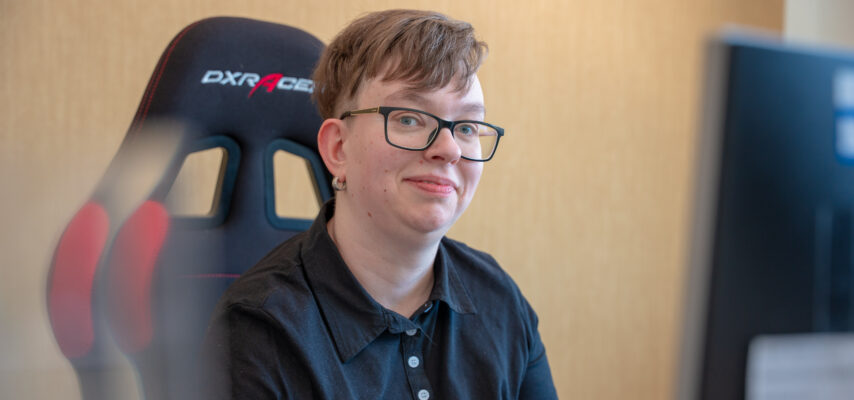 Person mit kurzen Haaren und Brille lächelt leicht vor einem Gaming-Stuhl, im Vordergrund unscharfer Monitor.