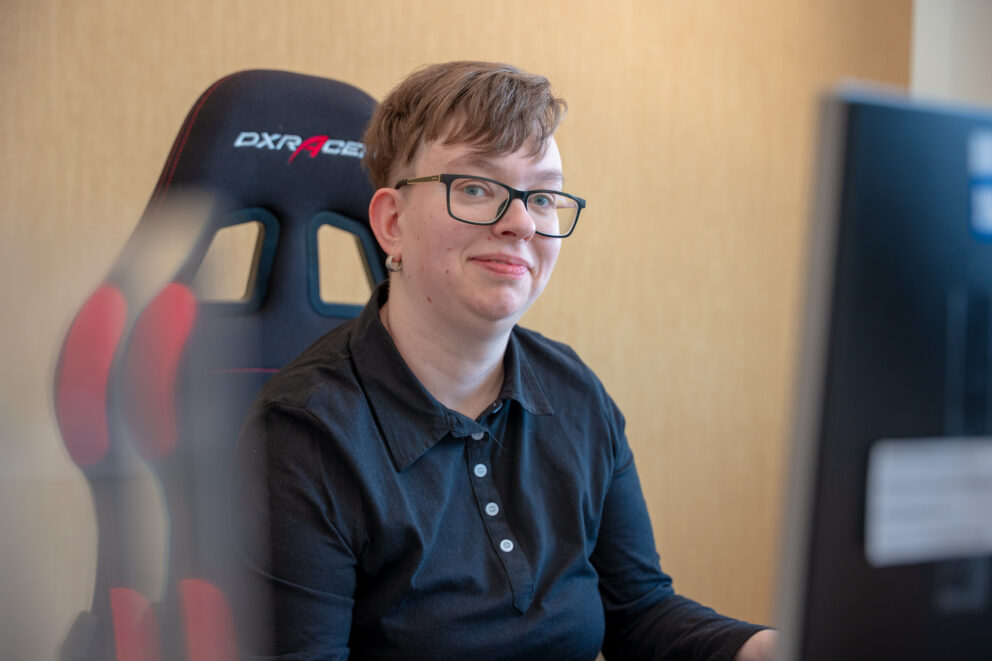 Person mit kurzen Haaren und Brille lächelt leicht vor einem Gaming-Stuhl, im Vordergrund unscharfer Monitor.