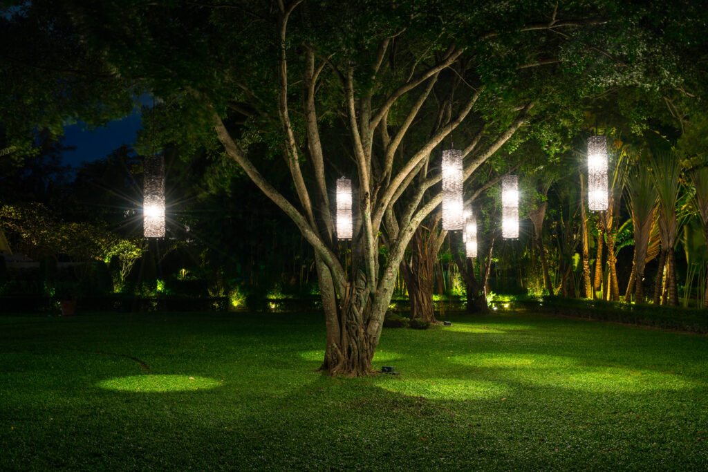 Großer Baum in einem gepflegten Garten bei Nacht, an dessen Ästen mehrere leuchtende zylindrische Laternen hängen und Rasen und Pflanzen beleuchten.