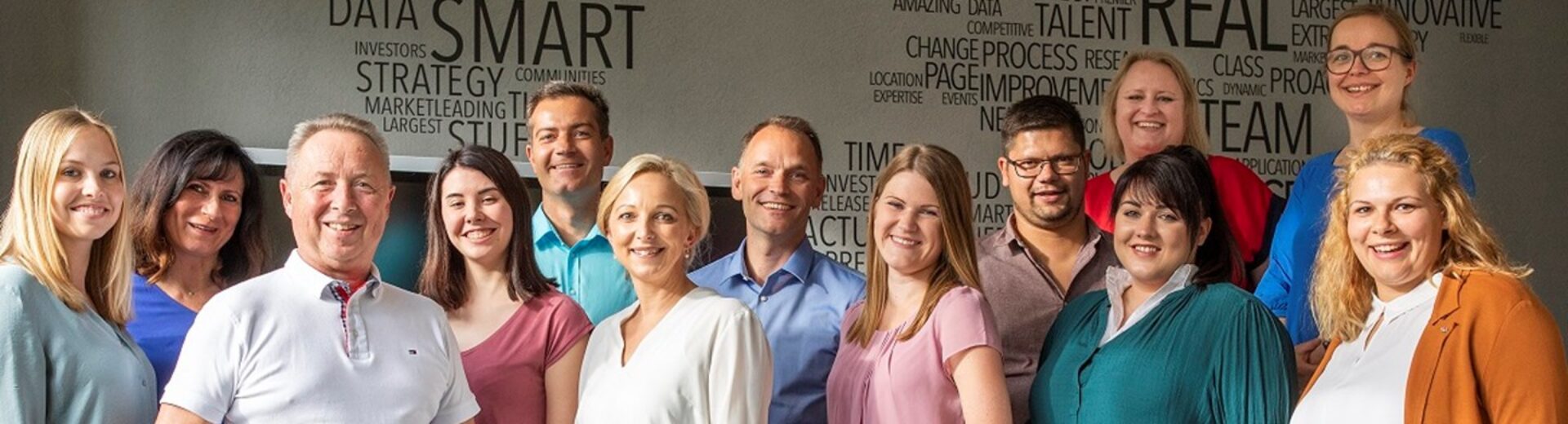 Gruppenfoto von zwölf lächelnden Büroangestellten in legerer Kleidung vor einer Wand mit Wortwolke (Schlagwörter wie SMART, REAL, DATA).