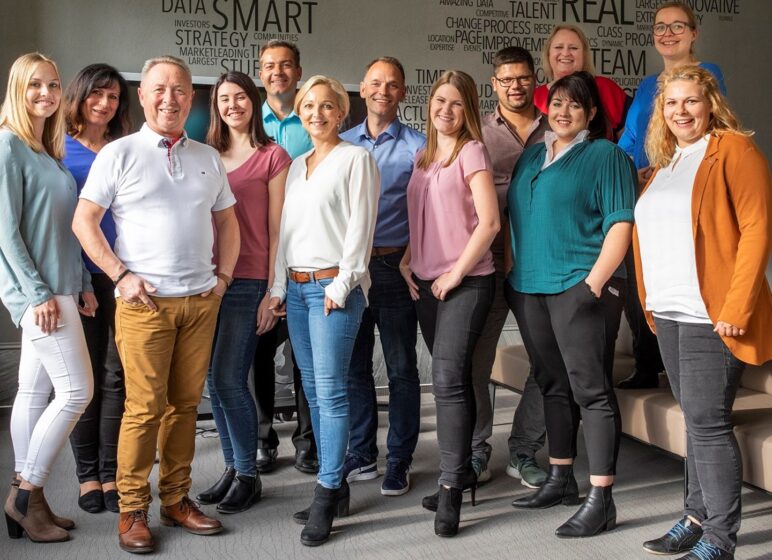 Gruppenfoto von zwölf lächelnden Büroangestellten in legerer Kleidung vor einer Wand mit Wortwolke (Schlagwörter wie SMART, REAL, DATA).