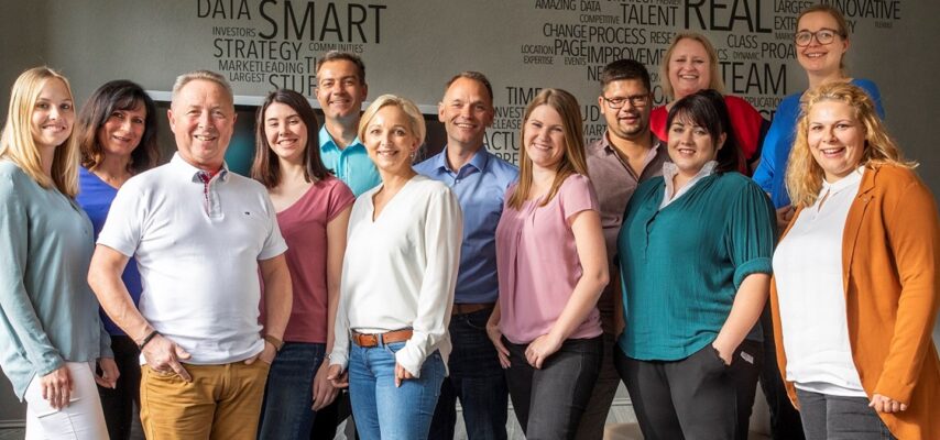 Gruppenfoto von zwölf lächelnden Büroangestellten in legerer Kleidung vor einer Wand mit Wortwolke (Schlagwörter wie SMART, REAL, DATA).