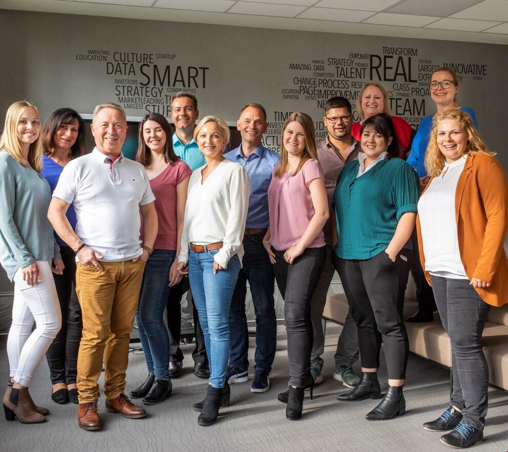 Gruppenfoto von zwölf lächelnden Büroangestellten in legerer Kleidung vor einer Wand mit Wortwolke (Schlagwörter wie SMART, REAL, DATA).