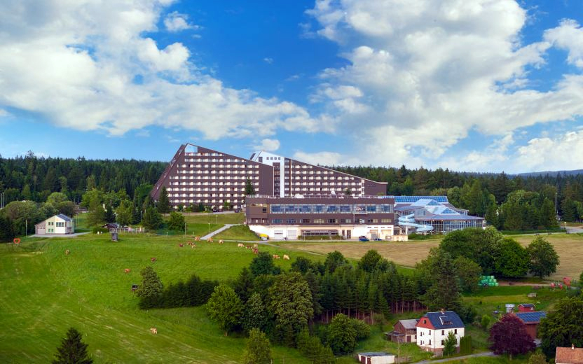 Großes, dreieckig zulaufendes Hotelgebäude am Waldrand über grünen Weiden mit vereinzelten Kühen und einigen Häusern im Vordergrund.