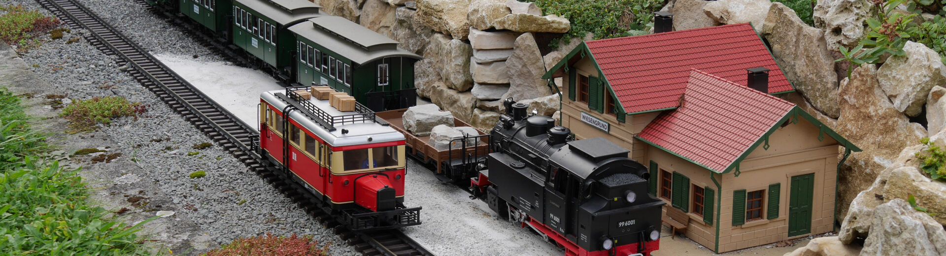 Gartenbahn mit schwarzer Dampflok, rotem Steuerwagen und grünen Personenwagen neben kleinem Bahnhofsgebäude und Felsenrabatte