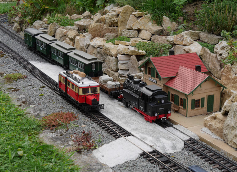 Gartenbahn mit schwarzer Dampflok, rotem Steuerwagen und grünen Personenwagen neben kleinem Bahnhofsgebäude und Felsenrabatte