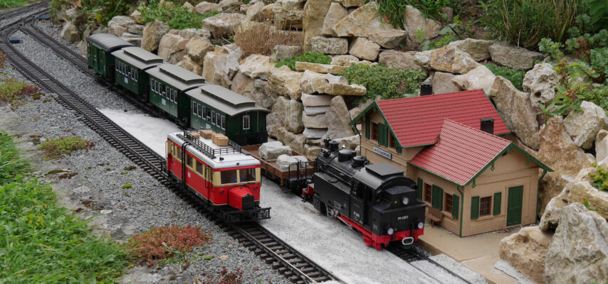 Gartenbahn mit schwarzer Dampflok, rotem Steuerwagen und grünen Personenwagen neben kleinem Bahnhofsgebäude und Felsenrabatte