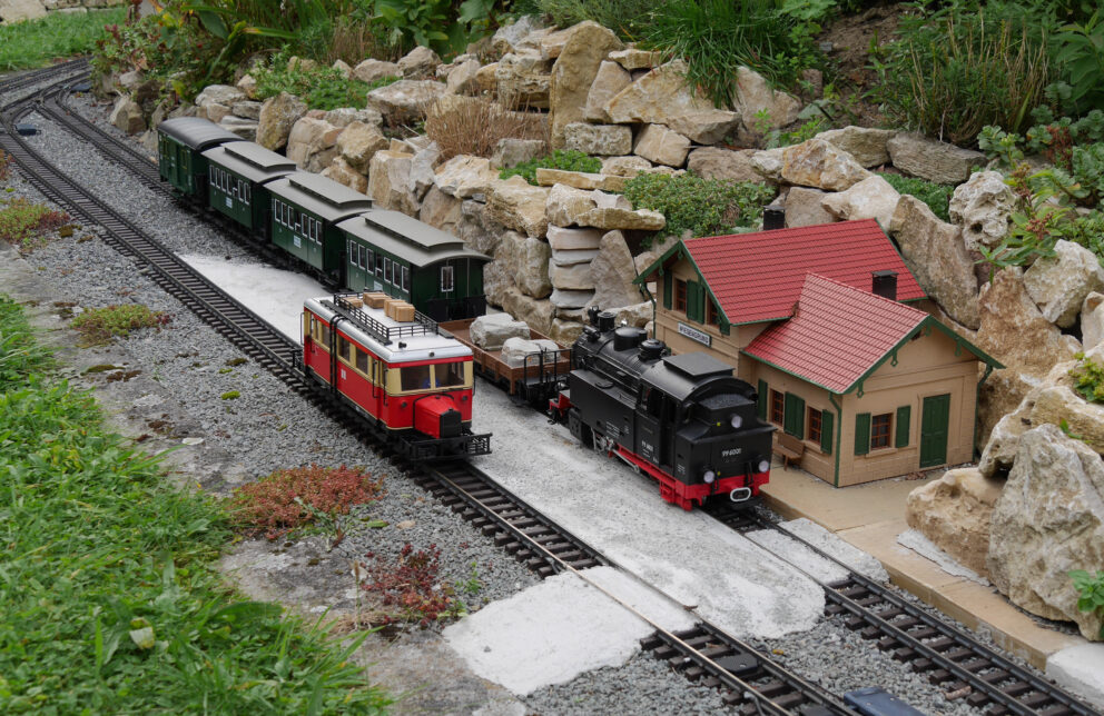 Gartenbahn mit schwarzer Dampflok, rotem Steuerwagen und grünen Personenwagen neben kleinem Bahnhofsgebäude und Felsenrabatte