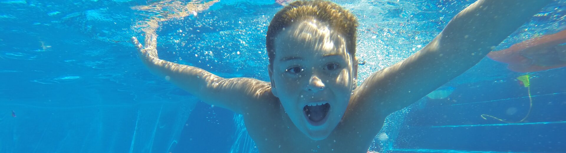 Junge taucht unter Wasser in einem hellblauen Pool, mit ausgestreckten Armen und offenem Mund, umgeben von Luftblasen.