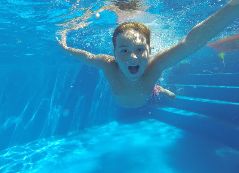 Junge taucht unter Wasser in einem hellblauen Pool, mit ausgestreckten Armen und offenem Mund, umgeben von Luftblasen.