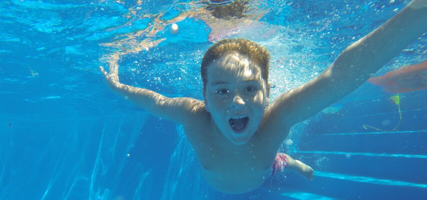Junge taucht unter Wasser in einem hellblauen Pool, mit ausgestreckten Armen und offenem Mund, umgeben von Luftblasen.