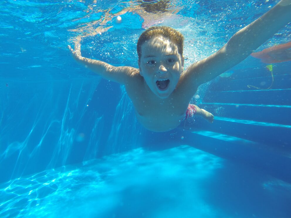 Junge taucht unter Wasser in einem hellblauen Pool, mit ausgestreckten Armen und offenem Mund, umgeben von Luftblasen.