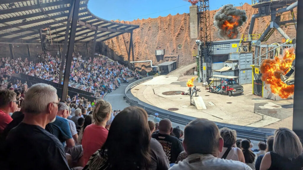 Publikum in überdachter Arena beobachtet Stuntshow mit Explosionen, Stuntperson auf Boden und Fahrzeug auf abgesperrter Aktionsfläche.