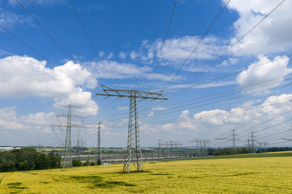 Hochspannungsmasten und -leitungen über weiten gelben Getreidefeldern unter blauem Himmel mit vereinzelten Wolken.