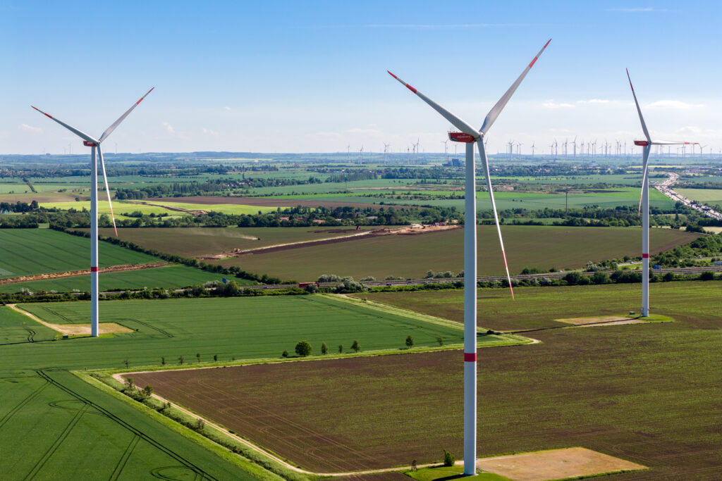 Drei große Windräder mit rot markierten Blattspitzen über weitläufigen grünen Feldern und weiteren Windrädern am Horizont.