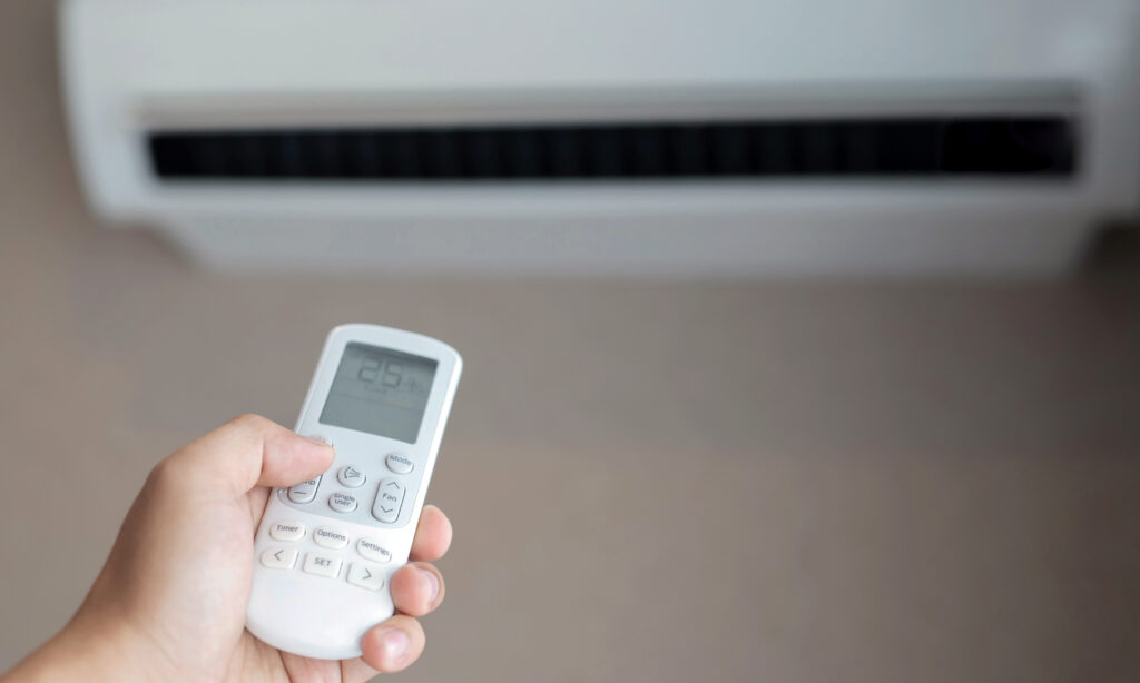Hand hält Fernbedienung, zielt auf wandmontierte Klimaanlage; Display zeigt Temperatur, Lüftungsgitter im Hintergrund.
