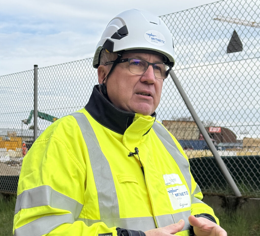 Mann mit Brille in weißem Schutzhelm und gelber Warnjacke vor einem Maschendrahtzaun auf Baustelle.