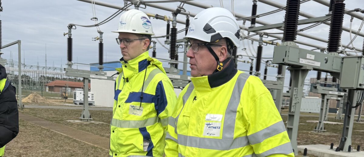 Zwei Personen in gelben Sicherheitsjacken und Schutzhelmen gehen durch eine Freiluft-Umspannanlage mit Hochspannungsisolatoren und Leitungsrohren im Hintergrund.