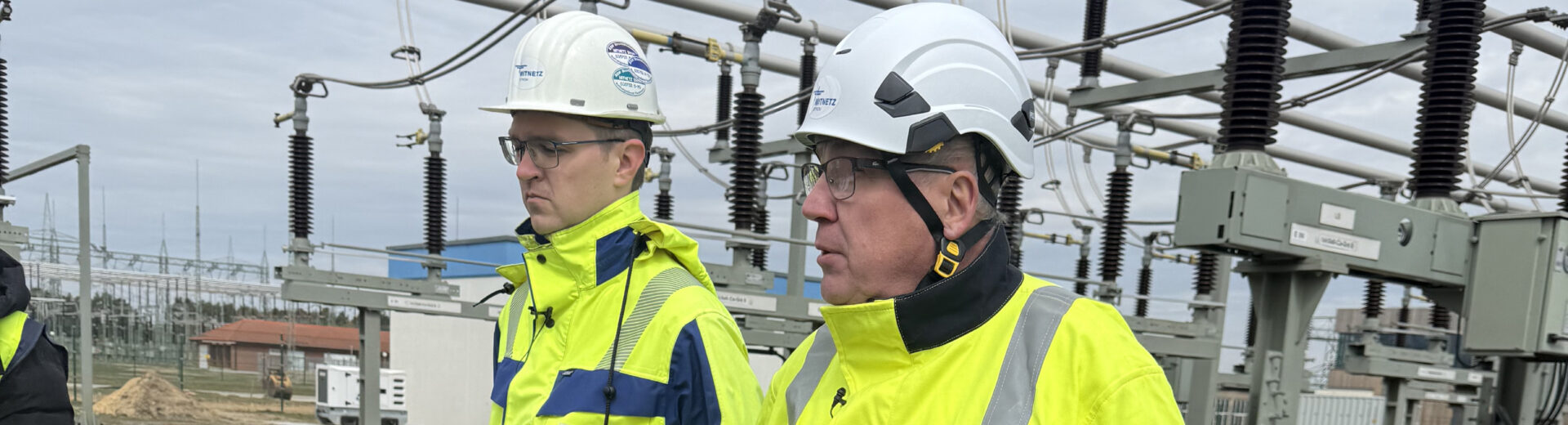 Zwei Personen in gelben Sicherheitsjacken und Schutzhelmen gehen durch eine Freiluft-Umspannanlage mit Hochspannungsisolatoren und Leitungsrohren im Hintergrund.
