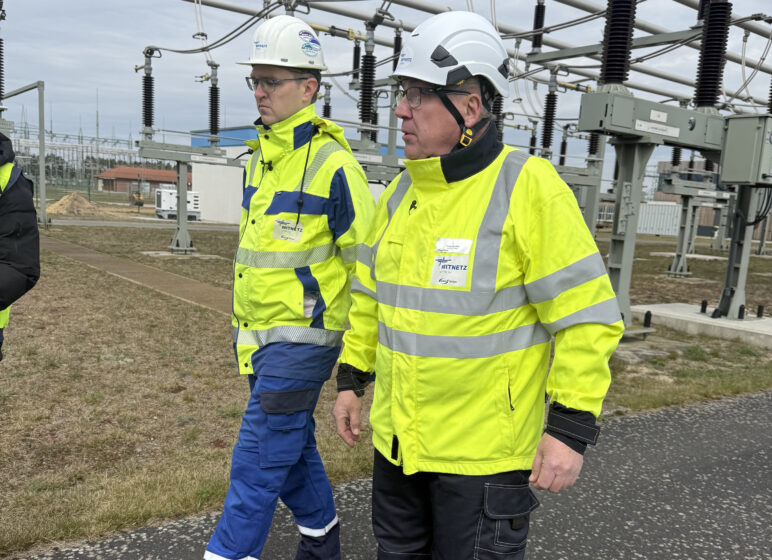 Zwei Personen in gelben Sicherheitsjacken und Schutzhelmen gehen durch eine Freiluft-Umspannanlage mit Hochspannungsisolatoren und Leitungsrohren im Hintergrund.