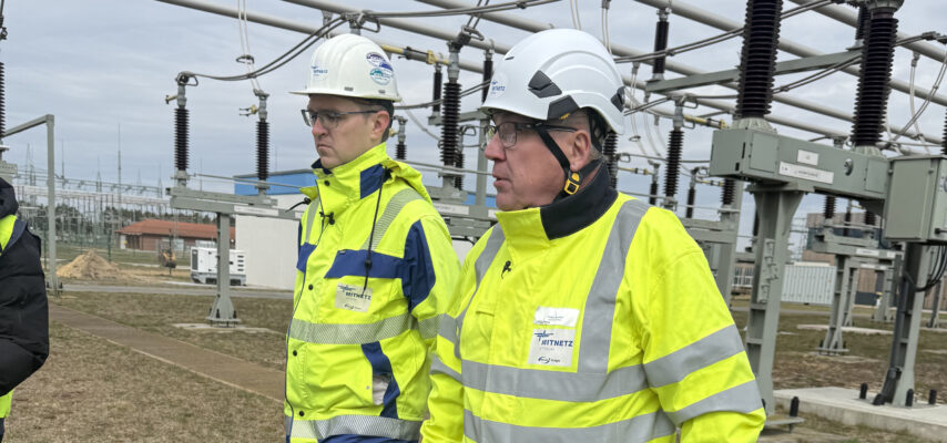 Zwei Personen in gelben Sicherheitsjacken und Schutzhelmen gehen durch eine Freiluft-Umspannanlage mit Hochspannungsisolatoren und Leitungsrohren im Hintergrund.