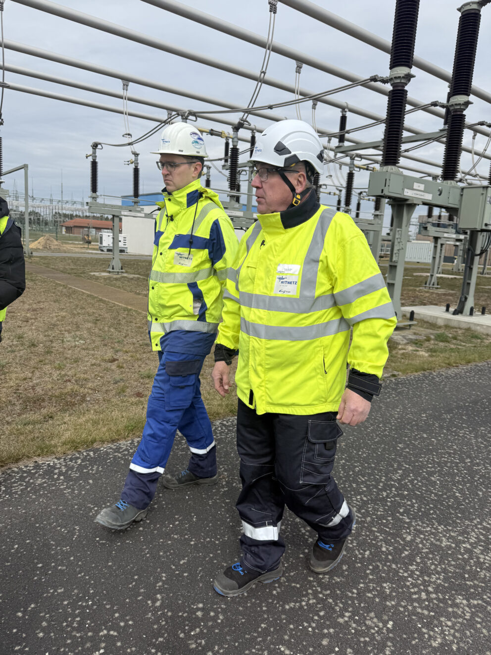 Zwei Personen in gelben Sicherheitsjacken und Schutzhelmen gehen durch eine Freiluft-Umspannanlage mit Hochspannungsisolatoren und Leitungsrohren im Hintergrund.
