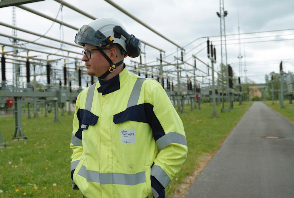 Elektriker in gelber Schutzjacke und Helm steht mit verschränkten Händen auf einem Umspannwerksgelände und blickt nach links.