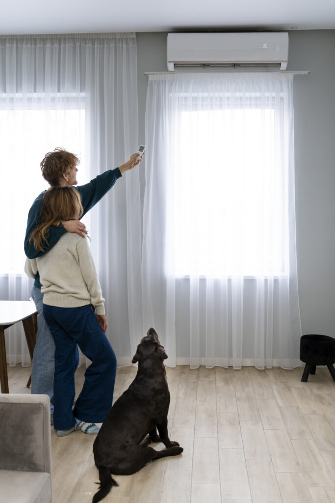 Paar steht umarmt vor hellem Fenster und richtet mit Fernbedienung die Wand-Klimaanlage, Hund sitzt daneben.
