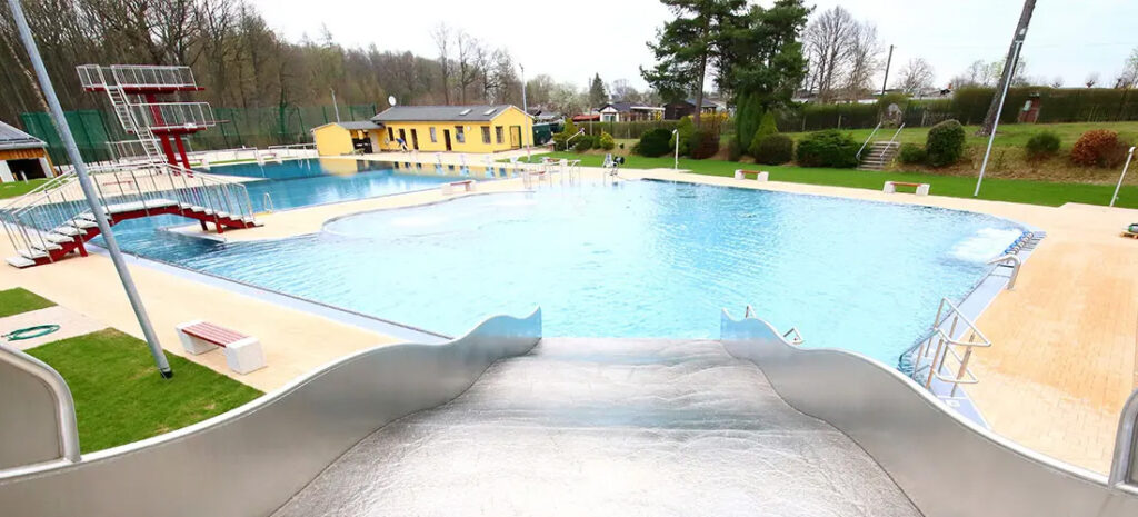 Blick vom oberen Ende einer Rutsche auf ein großes Freibad mit blauer Wasserfläche, Sprungturm links und gelbem Betriebsgebäude im Hintergrund