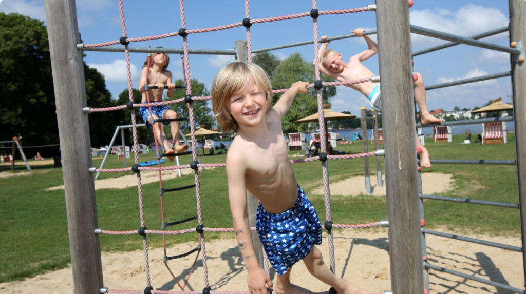 Lachender Junge in blau-weißen Stern-Badeshorts klettert an einem Seilnetz im Sandspielbereich, im Hintergrund zwei weitere spielende Kinder und See.