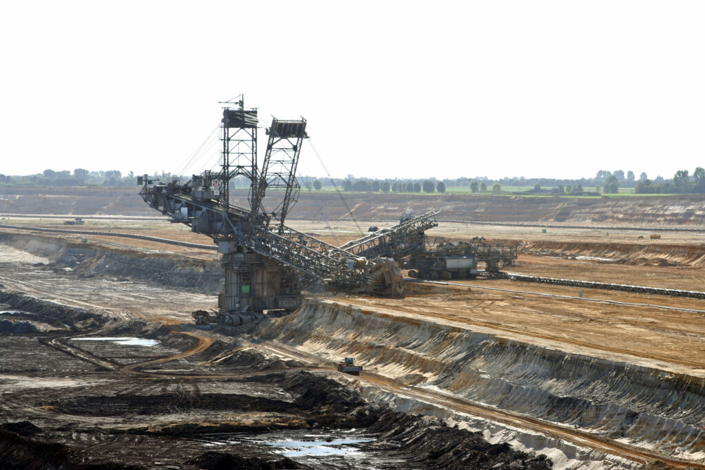 Große Schaufelradmaschine und Förderbänder in einem offenen Braunkohle-Tagebau vor weitläufigen Abraumflächen und Baumreihe am Horizont