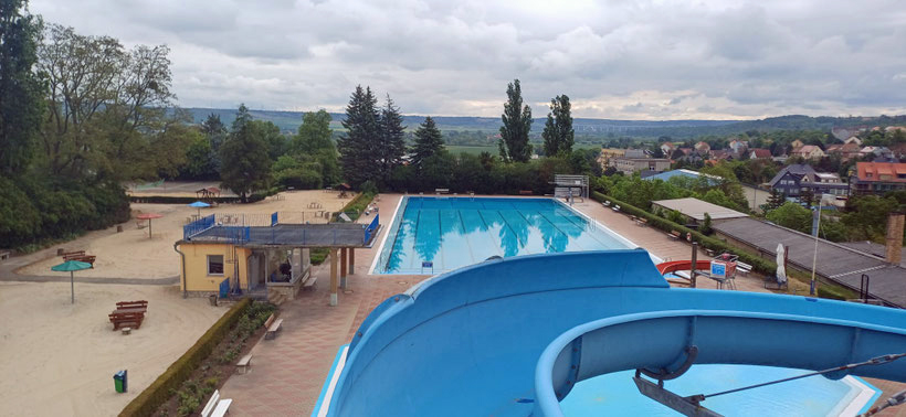 Freibad mit großem rechteckigem Schwimmbecken, blauer Wasserrutsche im Vordergrund und Sandspielbereich links, Bäume und Häuser im Hintergrund.