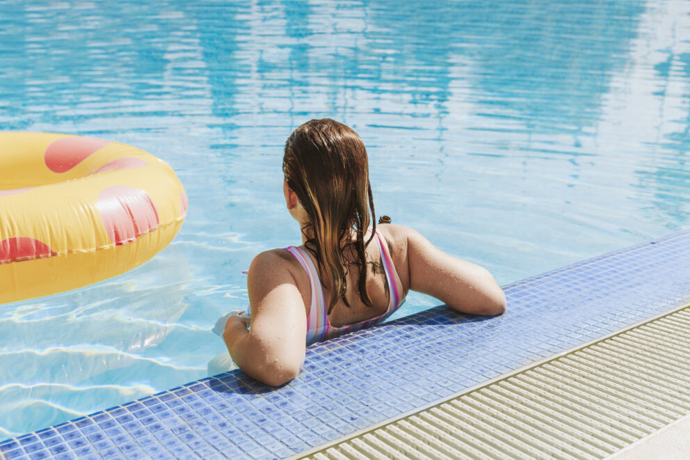 Frau von hinten in buntem Badeanzug lehnt am Beckenrand eines klaren blauen Schwimmbeckens, links ein gelb-rosa Schwimmring.