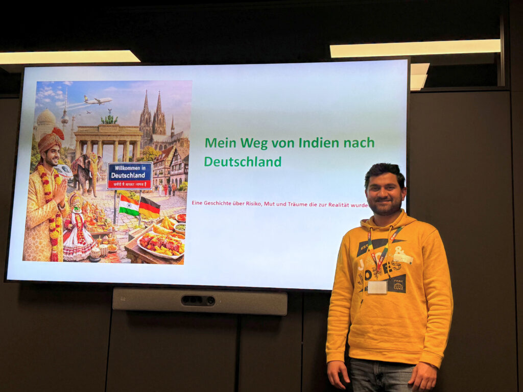Mann in gelbem Hoodie steht vor Präsentationsbild mit Titel „Mein Weg von Indien nach Deutschland“ und buntem Collage-Bild links.