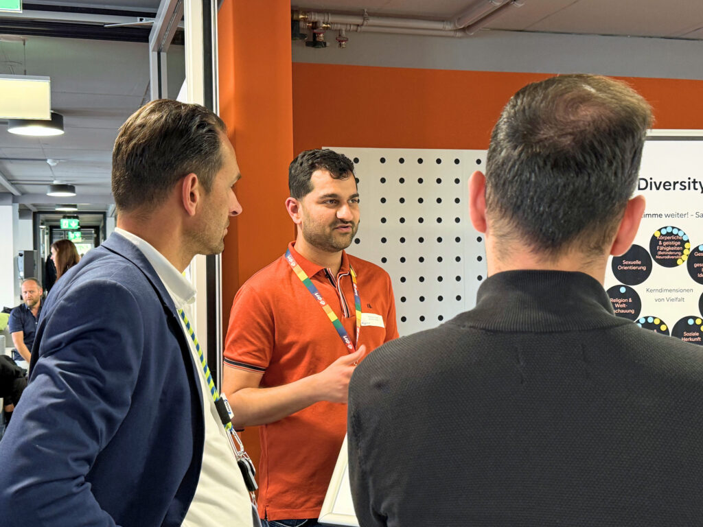 Drei Männer in Geschäftskleidung unterhalten sich vor einer orange-weißen Messestandwand; im Mittelpunkt ein Mann mit rotem Polo und Namensschild.