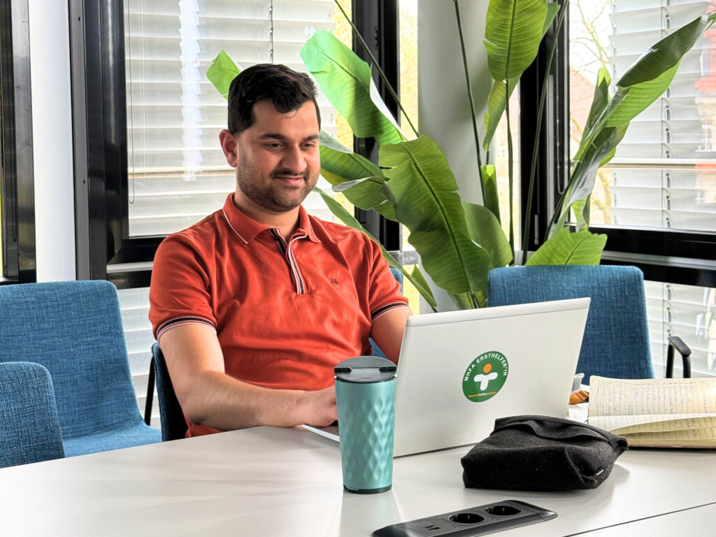 Mann in orange Poloshirt arbeitet an einem Laptop in einem hellen Büro, mit grüner Thermotasse und großen Zimmerpflanzen im Hintergrund.